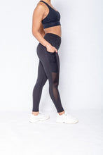 Load image into Gallery viewer, Shakolo crossover bra in black and mid waist leggings in black side view model stepping forward and one arm in pocket